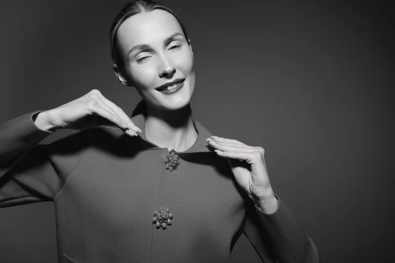 Smiling caucasian woman with pale skin and dark hair adjusts sweater collar, posing confidently on dark studio background. Concept of fashion and elegance.