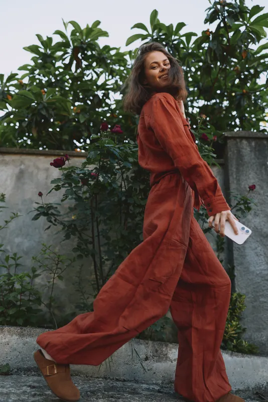 Woman lifestyle portrait of a woman in burnt orange jumpsuit walking outdoors, showcasing casual confident mood, candid street style against lush greenery, old stone wall, soft breeze, urban mood