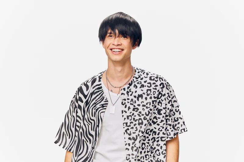 Young man smiling wearing a half zebra, half leopard patterned shirt and layered necklaces, standing against a white background with modern casual style.