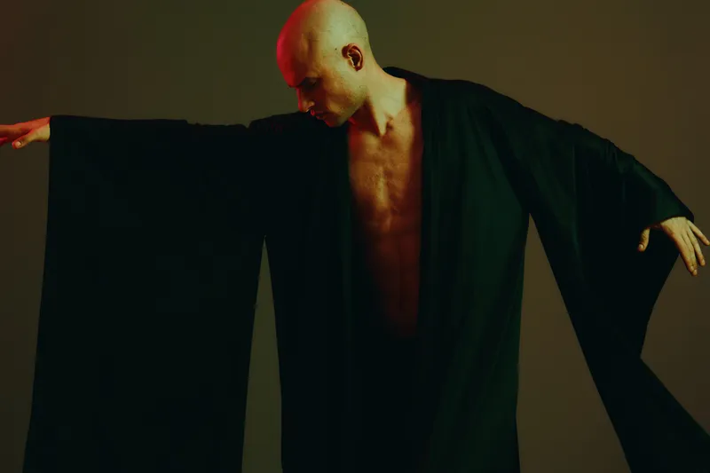 A striking man in a flowing black robe, showcasing his strong physique with an artistic pose, captured in a dimly lit setting. This black and white image emphasizes elegance.