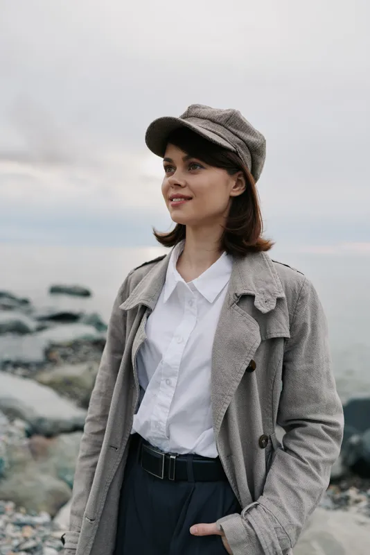 Young woman wearing a tweed coat and matching cap stands outdoors near rocky shore. She smiles softly with ocean and cloudy sky in the background, casual autumn fashion concept.