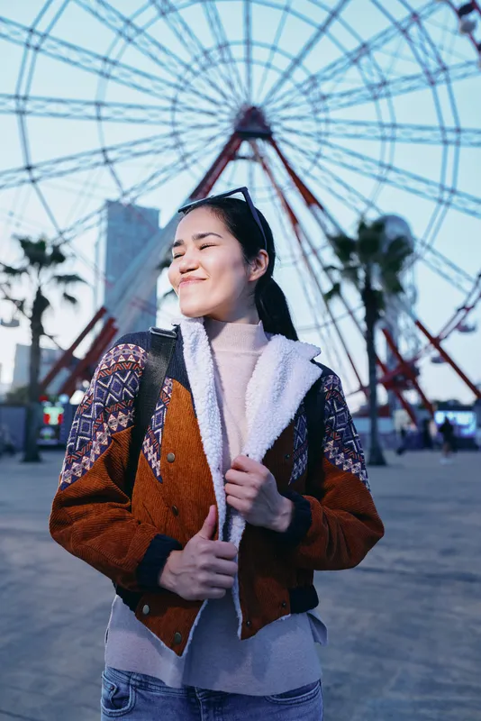 Smiling woman in a cozy jacket strolls through an outdoor amusement park with a large ferris wheel in the background, capturing carefree style, autumn light, and joyful adventure