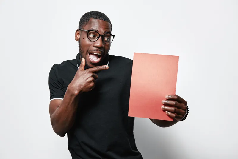 African American man with dark skin and glasses holding red paper and pointing at it with excitement on white background. Casual clothing and expressive face with open mouth.