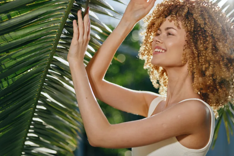 smiling woman with curly hair enjoying tropical outdoors, natural sunlight, green palm leaves, relaxed mood, summer lifestyle, bright warm colors