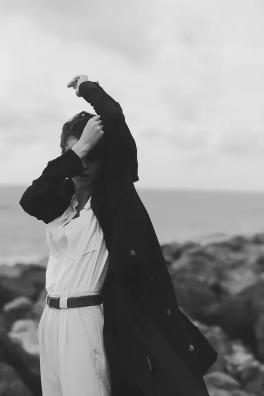 Fashionable coat dances in coastal wind as a figure on the shore adjusts a jacket, black and white mood, moody lighting, dynamic movement, seaside style
