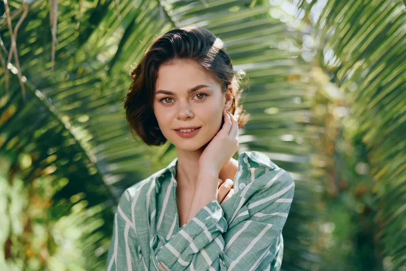 Portrait of a young woman in a striped shirt, bathed in warm natural light, standing among lush palm leaves with a relaxed, inviting expression.