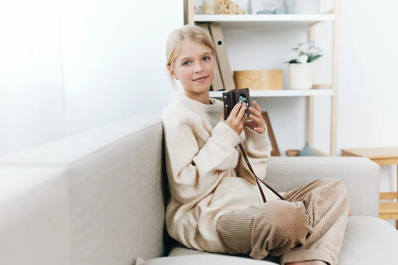 Young girl sits on a light sofa in a cozy living space, holding a compact camera and smiling softly, capturing a casual moment of creative play and relaxed photography.