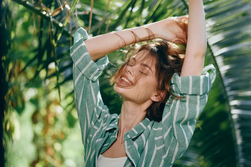 A joyful woman stands among lush tropical greenery with arms raised, smiling under warm sunlight. Casual striped shirt conveys carefree summer energy.
