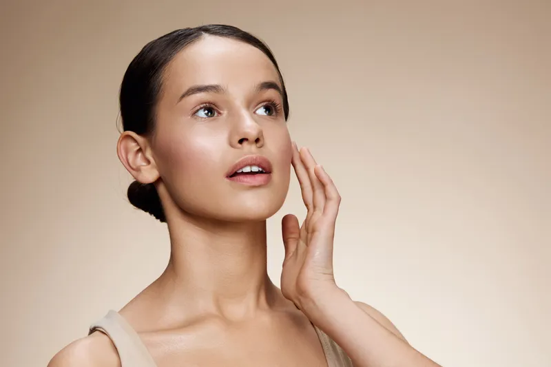 Portrait of an elegant young woman with smooth skin and a natural look, gently caressing her face, emphasizing beauty and skincare concepts in soft tones