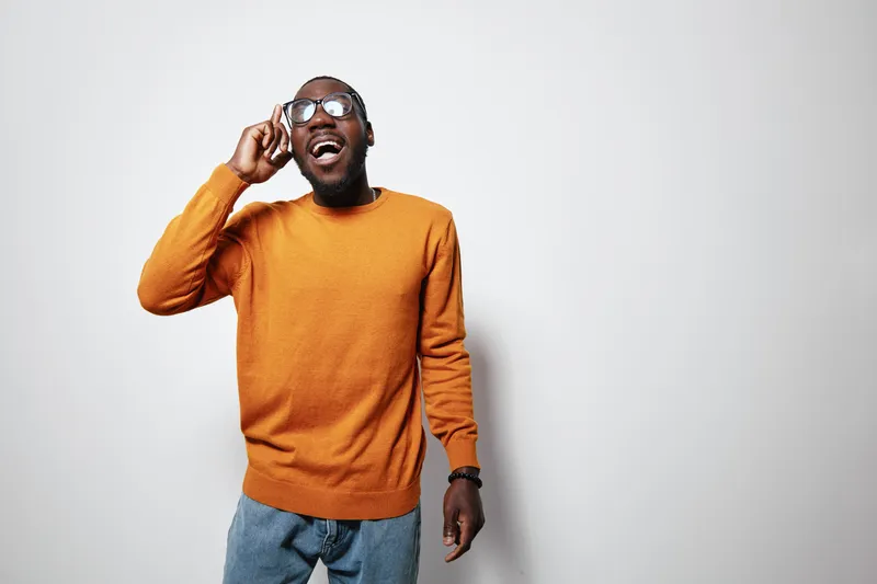African American man with dark skin wearing orange sweater and glasses talking on smartphone smiling isolated on white background. Casual style and happy expression.