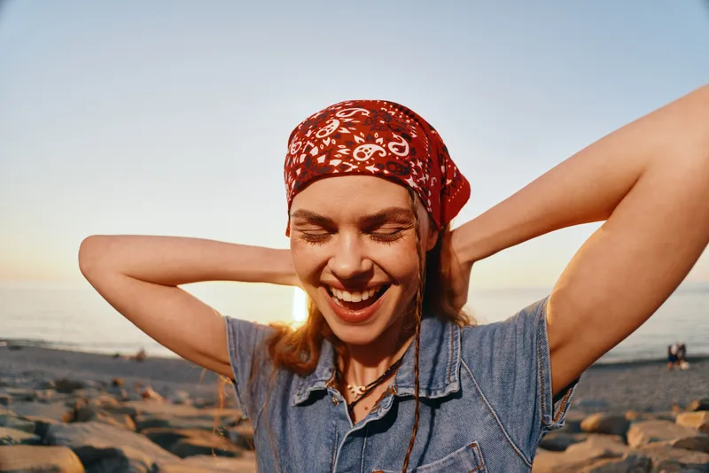 smiling woman with red bandana enjoying sunset on rocky beach in casual denim vest and natural warm light