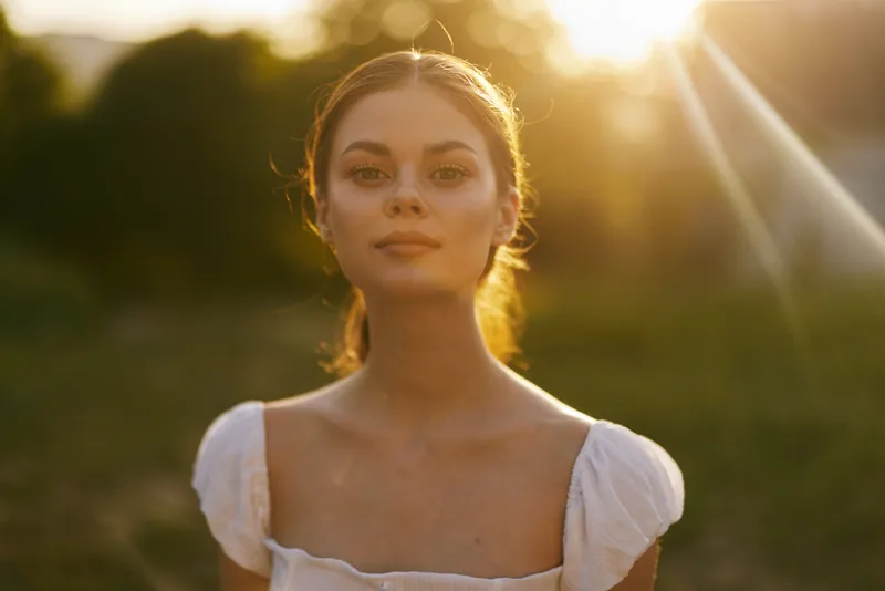 A serene portrait of a young woman in a white dress, glowing in the golden sunset, radiating warmth and confidence in a natural outdoor setting