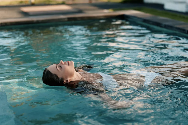 Relaxed woman floats on her back in a tranquil pool, enjoying sunlit water and a peaceful moment, minimal outdoor setting, serenity and self care, carefree summer vibe.