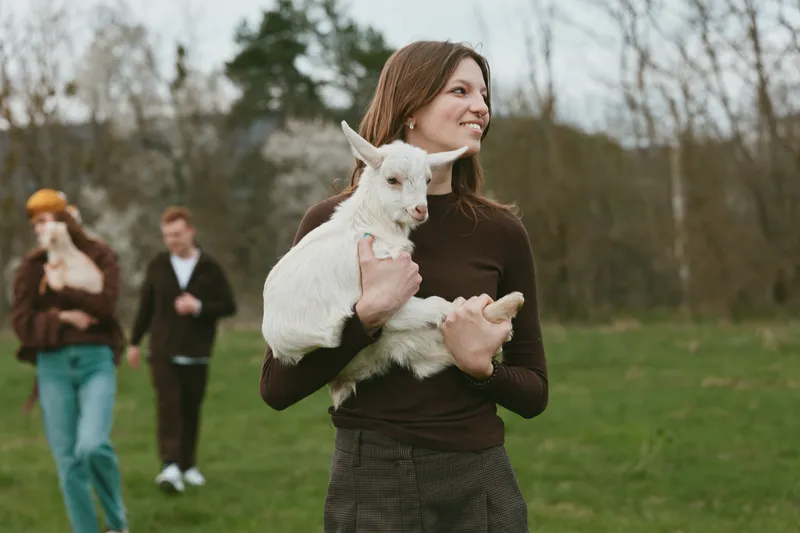 A woman in a dark sweater holds a white goat kid in a grassy field, with two people in the background. A candid lifestyle moment highlighting animal companionship and rural calm.