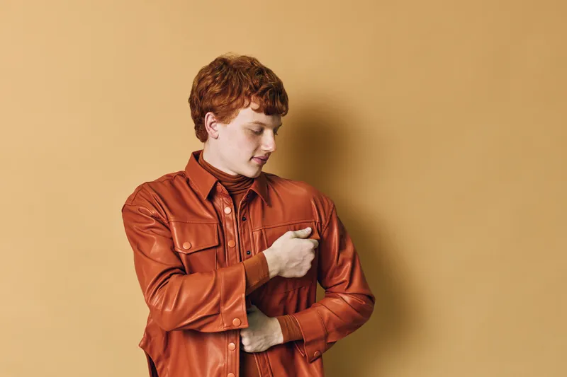 Redhead young man in leather jacket posing with hand on chest on beige background. Casual fashion portrait of male model with short hair and closed eyes, serene expression.