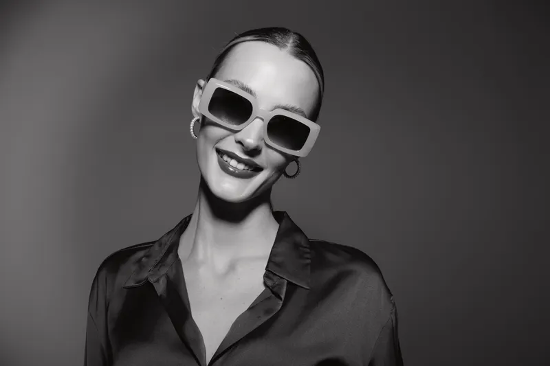 Smiling Caucasian woman with pale skin wearing oversized sunglasses and hoop earrings poses confidently in a dark satin blouse on grey background. Fashion and style concept.
