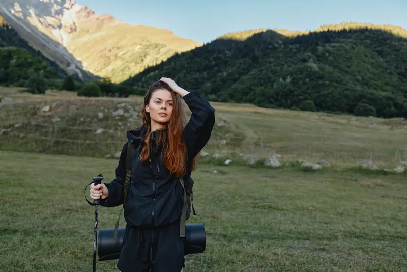 A confident female hiker stands in a wide alpine meadow, equipped with a trekking pole and a backpack. She smiles softly, soaking in the tranquil mountain landscape.