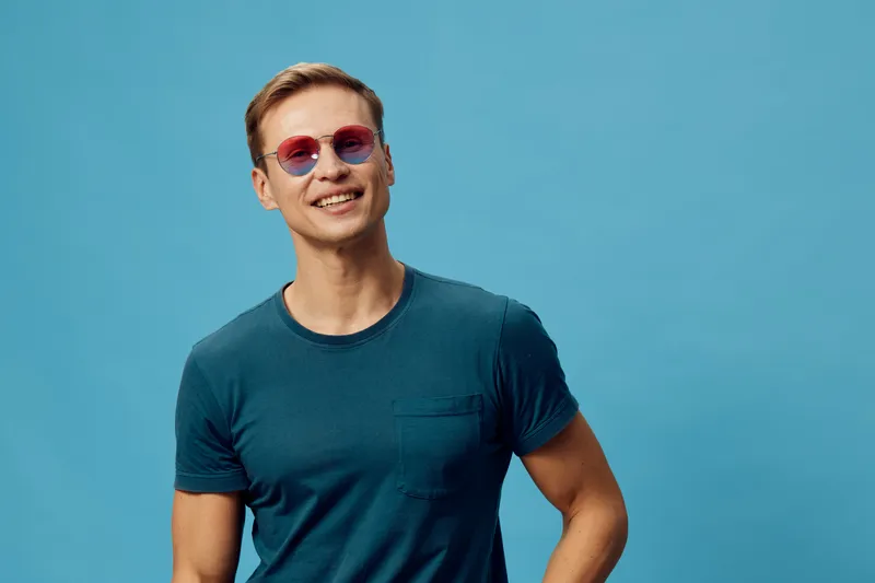 Smiling young caucasian man wear blue t-shirt and pink sunglasses isolated on blue background casual style digital nomad concept