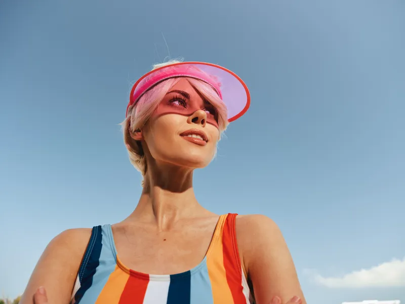 A confident woman in a bright striped swimsuit and pink visor stands outdoors against a clear blue sky, radiating summer energy and carefree confidence.