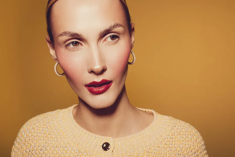 Close-up portrait of a stylish woman with red lipstick and gold hoop earrings wearing a textured beige sweater. Warm studio lighting highlights her smooth skin and confident expression.