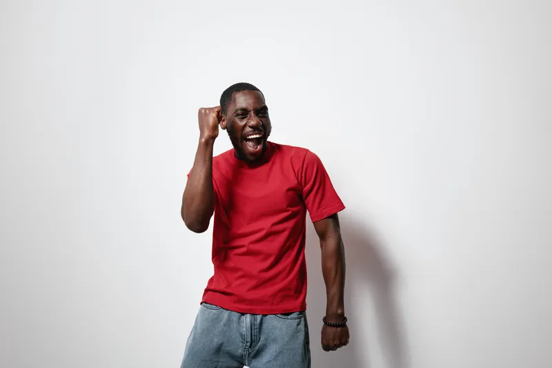 African American man with dark skin wearing red t shirt and jeans celebrating with fist pump and open mouth against white background in studio. Excited, happy, casual style.