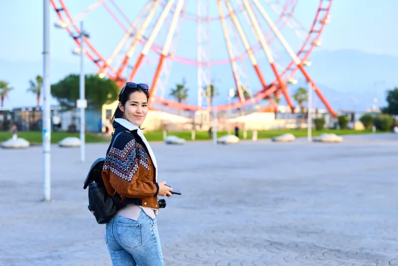 Woman with camera in outdoor amusement park beside a large ferris wheel, casual jacket, backpack, exploring and smiling as the scene captures travel and leisure moments