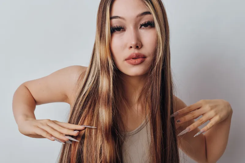 Young woman with long highlighted straight hair posing with hands near hair against white background. Fashion, beauty, female portrait with natural makeup and casual top.