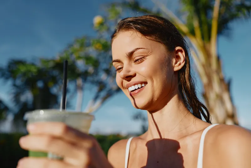 Outdoor woman smiling, holding a drink in a sunny summer setting, highlighting casual lifestyle and hydration in a bright portrait that conveys simplicity and health.