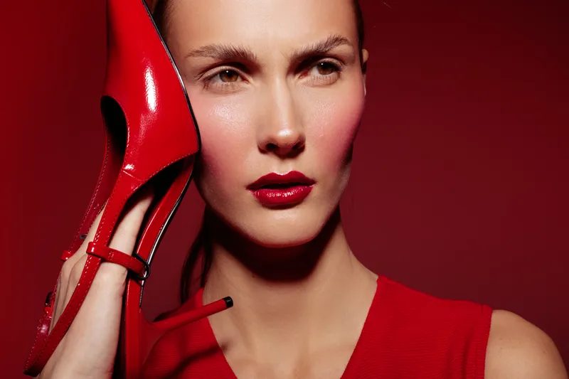 Close-up of a woman with red lipstick and red dress holding a red high heel shoe near her face. The intense red background creates a bold fashion and beauty concept with strong color contrast.