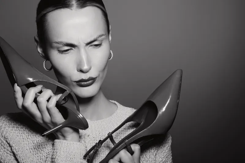Caucasian woman with light skin holds high heel shoes near face, showing confused expression. Concept for fashion, footwear, and style doubts.