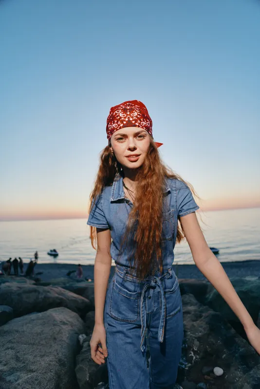Lifestyle woman in modern boho-western denim street style with a red bandana stands by the seaside during warm sunset film color tones.