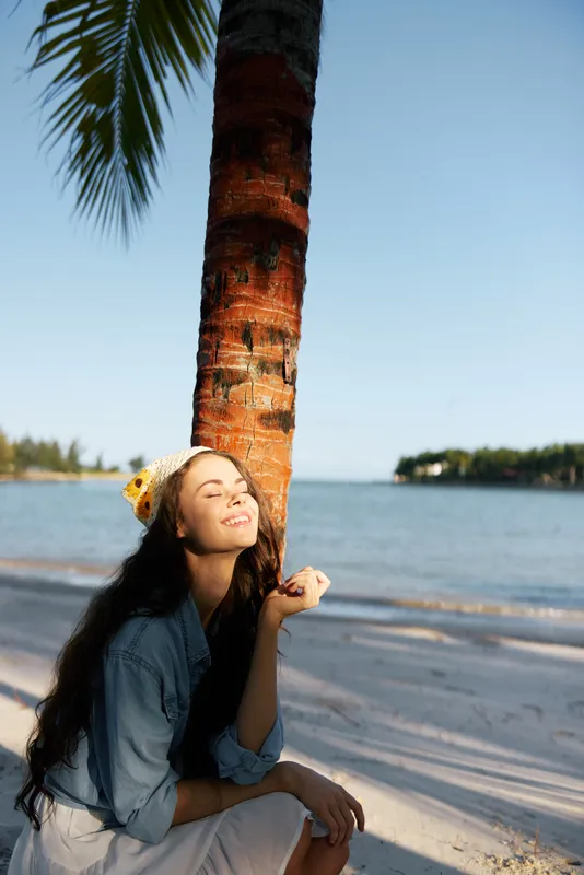 A woman sits by a palm tree on a sunny beach, eyes closed, smiling softly, denim jacket, hair flowing, warm light, calm breeze, tranquil seaside moment, carefree.