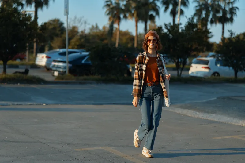 Woman walking in a casual urban scene, smiling with tote and beanie in warm golden hour glow. Authenticity, candid lifestyle, mindful living and emotional storytelling in modern street portrait.