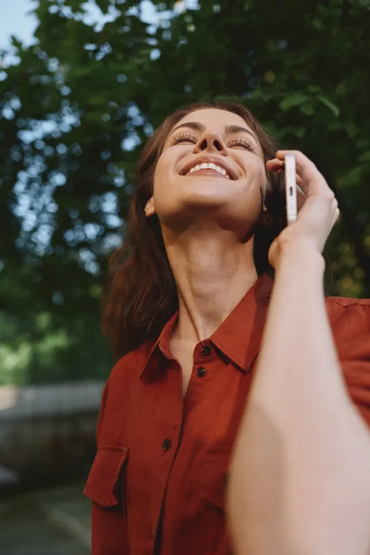 Young woman with dark hair talks on a smartphone outdoors, smiling warmly as she stands beneath lush green trees on a sunny day, conveying casual daily life and connection
