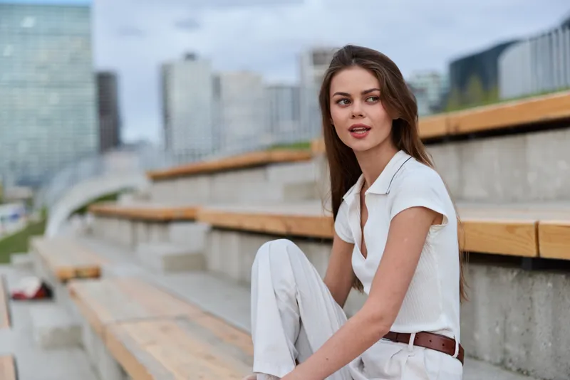 Young woman in white casual outfit sitting on urban wooden bleachers with thoughtful expression and modern cityscape background outdoor