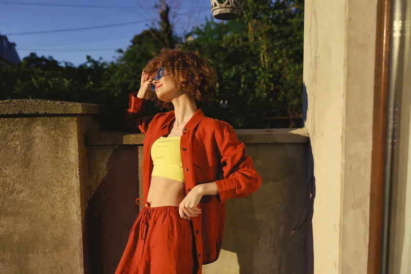 A stylish woman in a red outfit stands on a sunlit terrace, shielding eyes with her hand. Bright outdoor scene highlighting casual fashion and confident lifestyle