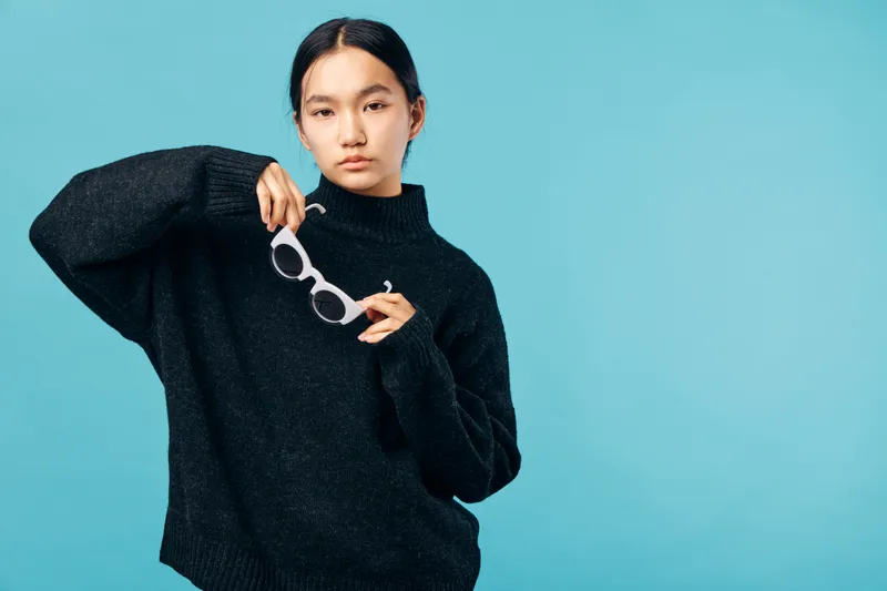 Young woman with dark hair wearing oversized black sweater holding white sunglasses against blue background. Casual fashion portrait with minimal makeup and neutral expression.