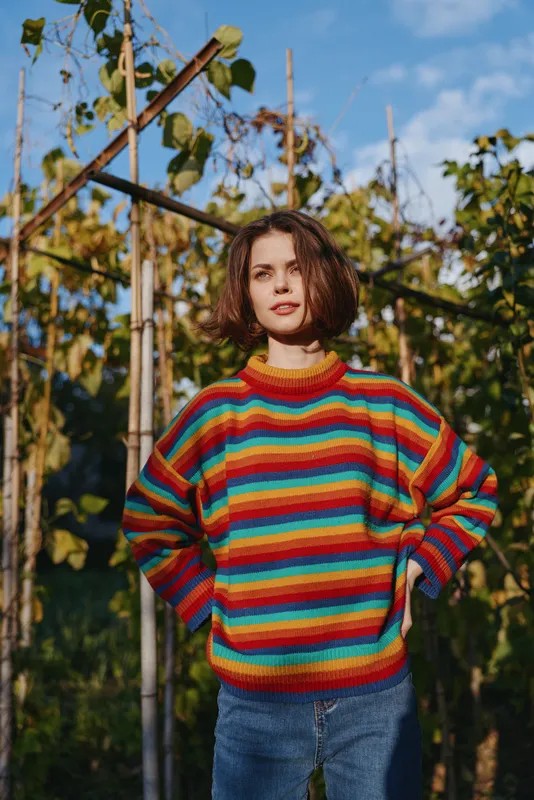 Woman portrait outdoors in rainbow sweater and jeans, casual full-length pose among plants and vines, short bob haircut and hands on hips, confident natural fashion in sunlight.