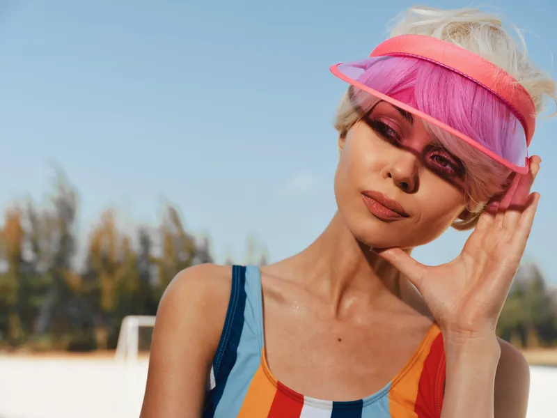 Bright portrait of a young woman wearing a pink visor and colorful striped swimsuit, sunlit face and relaxed pose against a clear beach backdrop, capturing vibrant summer energy and stylish outdoor