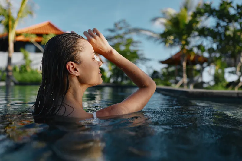 Woman swims in a bright outdoor pool at a tropical resort, water slicked hair, hand shielding eyes from the sun, relaxed smile, carefree summer vibe, vacation scene, tranquil moment, lifestyle.