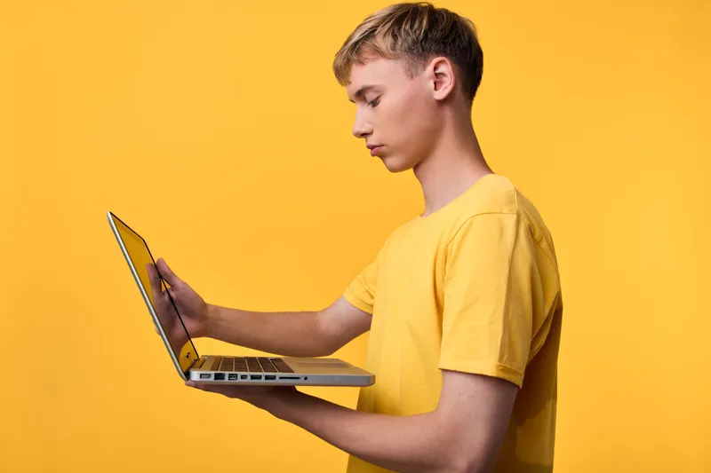 Young man in a yellow t shirt holds a laptop with one hand and taps the screen with the other, against a vivid yellow backdrop, illustrating modern technology, learning and digital work in action