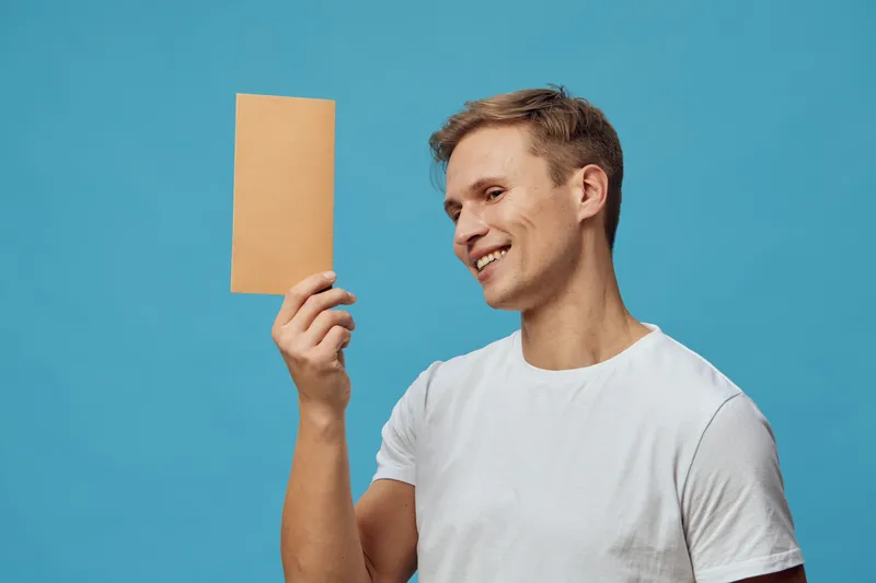 Smiling man with short hair in casual white T-shirt holding blank cardboard sign against blue background. Studio portrait. People lifestyle concept emphasizing communication, expression, advertising, 