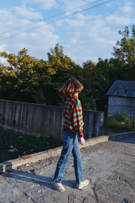 Woman walking in countryside wearing a colorful striped sweater, jeans and sneakers. Young adult with bob haircut, casual outfit, relaxed travel lifestyle in rural afternoon.