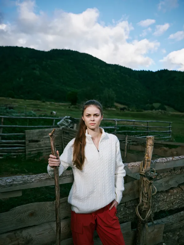 Woman outdoors in a white sweater and red pants stands beside a wooden fence with a staff in a rural countryside landscape under a blue sky and rolling hills
