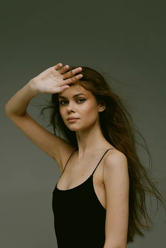 young woman shielding eyes with hand against dark background in black dress