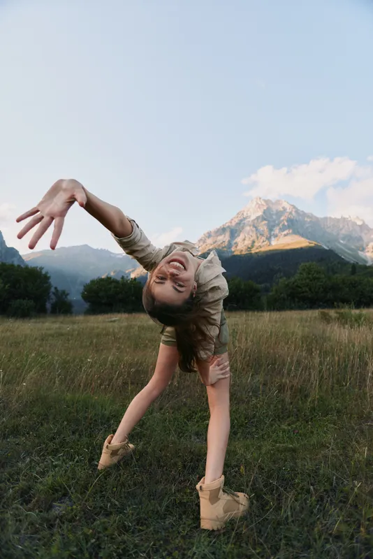 Playful woman performing a backbend in a sunlit field with mountains in the background, joyful energy, casual outfit, natural colors, carefree outdoor lifestyle