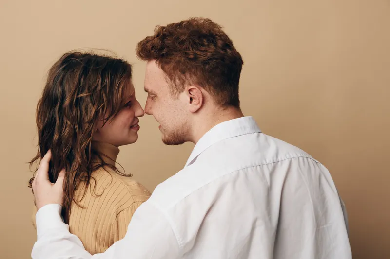 Young couple embraces closely, looking into each other’s eyes with affection. Warm beige background highlights intimate moment of love, connection, and tenderness between man and woman.