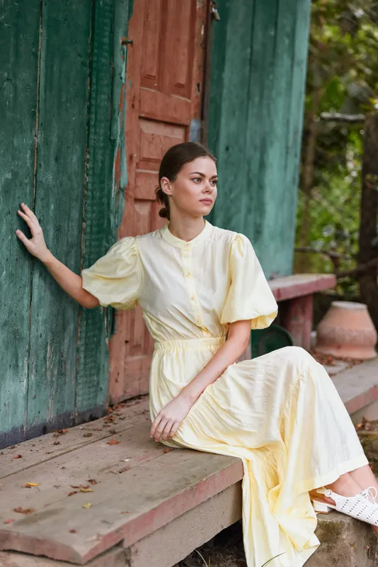 Elegant woman in a pale yellow dress sits on a wooden porch beside a green doorway in an outdoor setting, calm expression and soft light highlight style