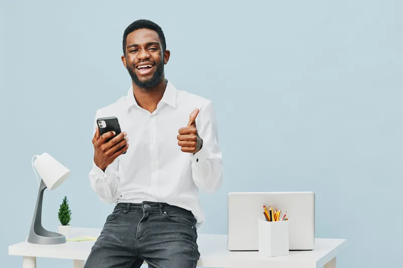 Smiling young African-American man in casual white shirt holding smartphone and giving thumbs-up gesture while sitting at modern white office desk with laptop, lamp, and potted plant on pastel blue ba