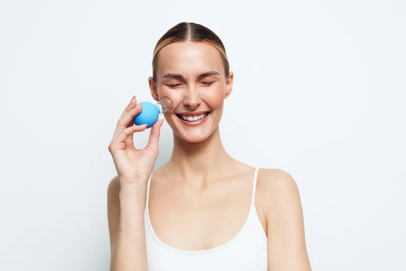 A smiling young woman with closed eyes holds a blue facial cleansing sponge near her cheek, demonstrating skincare routine and beauty care on a white background.
